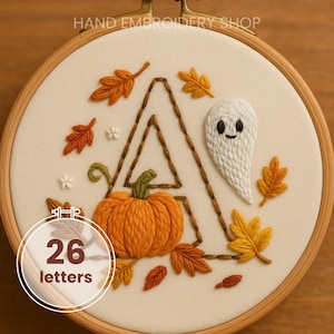 Puede incluir: Arte de aro bordado con la letra "A", una calabaza, un fantasma y hojas de otoño. El diseño incluye el texto "HAND EMBROIDERY SHOP" y "26 letters". El bordado está en un aro de madera.