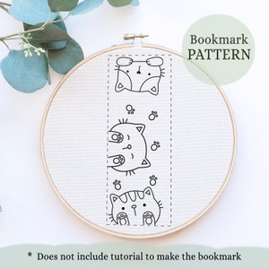 May include: A white embroidery hoop holds a canvas with a black and white cat bookmark pattern. The pattern features three cartoon cat faces and paw prints. Text reads "Bookmark PATTERN" and "Does not include tutorial to make the bookmark."