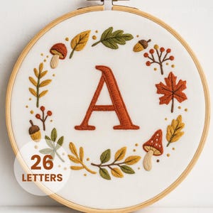 Puede incluir: Arte de aro bordado con la letra "A" en naranja quemado, rodeada por una corona de hojas de otoño, bellotas y setas en colores otoñales. El texto "26 LETTERS" está en un círculo blanco. El bordado está sobre tela blanca.