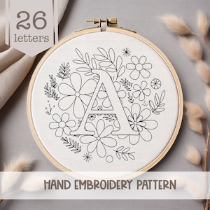 May include: A black and white hand embroidery pattern featuring the letter A surrounded by a wreath of flowers and leaves. The pattern is on a white fabric background and is labeled "HAND EMBROIDERY PATTERN".