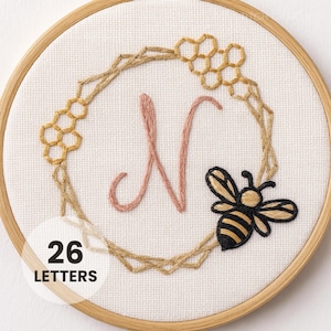 May include: Embroidered hoop art featuring a pink letter "N" surrounded by a stitched wreath with honeycomb and a bee. The design is on white fabric, set in a wooden hoop. The text "26 LETTERS" is at the bottom.