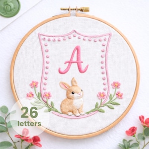 Può includere: Ricamo su telaio con la lettera "A" rosa all'interno di un disegno a scudo, con un coniglietto e dettagli floreali. Il ricamo è su tessuto bianco all'interno di un telaio di legno. Il testo "26 letters" è in basso a sinistra.