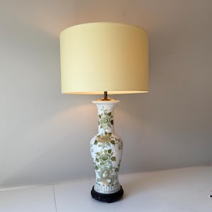 Puede incluir: Lámpara de mesa decorativa con pantalla color crema y base de cerámica blanca. La base está adornada con motivos florales verdes y descansa sobre una base de madera negra. La lámpara está diseñada para uso en interiores.