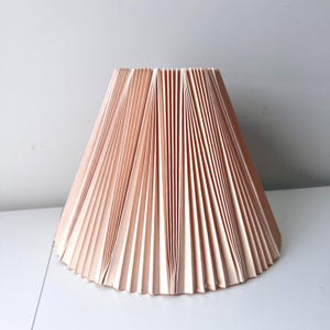 May include: A peach-colored lampshade with a pleated design. The lampshade has a conical shape, with vertical pleats creating a textured surface. The shade is sitting on a white surface.