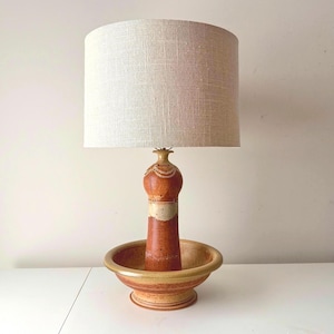 Puede incluir: Lámpara de mesa de cerámica con pantalla de lino beige. La base de la lámpara es de color marrón rojizo con una banda crema y un diseño decorativo. La lámpara se encuentra en un cuenco de cerámica a juego. La lámpara mide unos 76 cm de alto.
