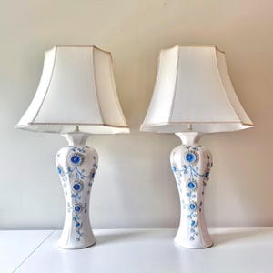 Puede incluir: Dos lámparas de mesa de cerámica blanca con detalles florales azules y blancos. Cada lámpara tiene una pantalla blanca con ribete dorado. Las lámparas están sobre una superficie blanca con un fondo neutro.