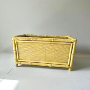 May include: A rectangular, light yellow bamboo planter. The planter has a slatted front and is constructed with bamboo poles. The corners are reinforced with decorative bamboo joints. The planter is elevated on short legs.