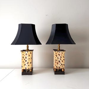 Puede incluir: Dos lámparas de mesa a juego con pantallas negras. Las lámparas tienen una base rectangular con un estampado moteado en beige y marrón. Las lámparas tienen un cuello color latón y una base marrón oscuro con esferas doradas.