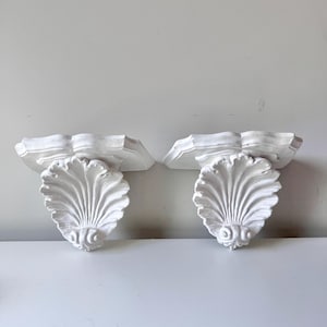 Puede incluir: Dos estantes de pared blancos en forma de concha. Cada estante presenta una parte superior festoneada y un diseño detallado en forma de abanico. Los estantes están hechos de un material sólido y están diseñados para uso decorativo o funcional.
