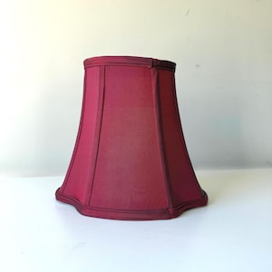 May include: A burgundy lampshade with a flared, trapezoidal shape. The shade has a pleated texture and is trimmed with a darker burgundy border. The lampshade is designed to sit on a lamp base and diffuse light.