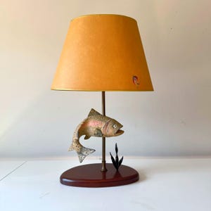Puede incluir: Una lámpara de mesa vintage con una escultura de pez. La lámpara tiene una pantalla naranja cálida con un borde dorado y un pequeño diseño. La base es de madera oscura ovalada, y el pez está pintado con colores naturales.