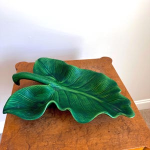 May include: A ceramic dish shaped like a large green leaf with a glossy finish. The dish features detailed embossed veins and a curved stem handle, showcasing a vibrant emerald green color with darker shading in the recessed areas of the leaf.