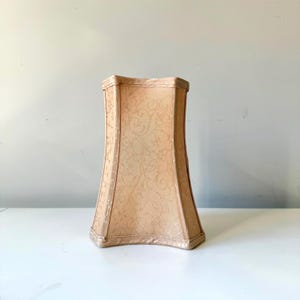 Może przedstawiać: Beżowy klosz lampy o zakrzywionym, prostokątnym kształcie. Klosz ma kwiatowy wzór i teksturowaną powierzchnię. Klosz stoi na białej powierzchni.