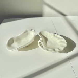 Puede incluir: Dos cuencos de cerámica blanquecina con forma de concha de ostra. Los cuencos tienen una superficie texturizada y un diseño naturalista. Las conchas están colocadas sobre una superficie blanca, con sombras.