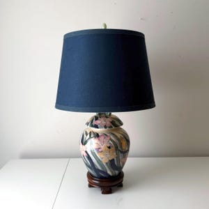 Puede incluir: Una pantalla de lámpara azul marino se asienta sobre una lámpara de cerámica con un diseño floral. La base de la lámpara tiene forma de jarrón con tapa, con motivos florales rosas, amarillos y verdes. La lámpara descansa sobre una base de madera marrón oscuro.