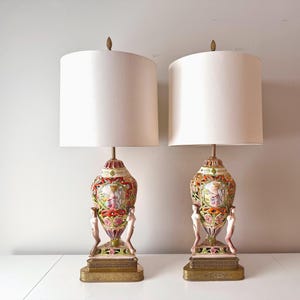 Puede incluir: Par de lámparas de mesa ornamentadas con pantallas color crema. Las lámparas presentan bases de cerámica detalladas y coloridas con figuras clásicas, diseños florales y detalles dorados. Cada lámpara tiene una base rectangular y un remate decorativo.