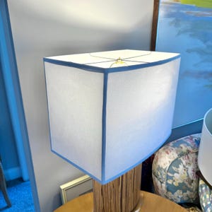 Puede incluir: Una pantalla de lámpara blanca con un borde azul claro se asienta sobre una base de lámpara de estilo bambú. La pantalla tiene forma rectangular con bordes curvos. La lámpara está sobre una mesa redonda de madera.