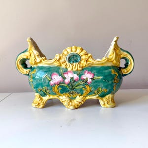 May include: A ceramic planter with a teal base, adorned with pink floral details and gold accents. The planter has a rectangular shape with ornate handles and feet, and is suitable for indoor use.