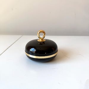 May include: A glossy, black, round container with a gold-colored band and a gold loop handle. The container is sitting on a white surface. The design is simple and elegant.