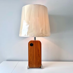 May include: A mid-century modern table lamp with a rectangular wooden base and a cream-colored lampshade. The base has a dark circular detail and sits on a square metal plate. The lampshade is covered in a translucent material.