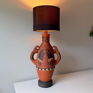 May include: A terracotta-colored ceramic table lamp with a black lampshade. The lamp base features a decorative design with two handles and a black base. The lampshade is cylindrical and made of a dark fabric.