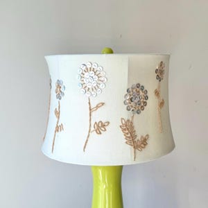 May include: A table lamp with a vibrant green base and a white lampshade. The lampshade is adorned with embroidered floral patterns, silver sequins, and brown thread. A small green sphere is positioned on top of the lampshade.