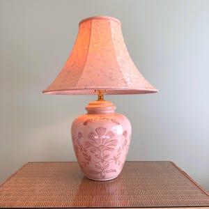May include: A table lamp with a pink ceramic base featuring a dark pink floral pattern and a matching pink fabric lampshade. The lamp emits a warm glow, highlighting the intricate botanical etchings on the rounded, bulbous ceramic body.