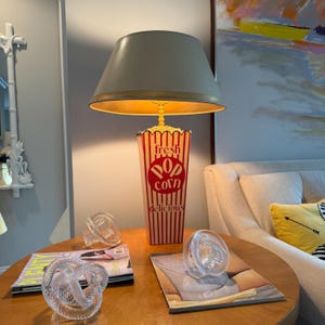 Puede incluir: Una lámpara novedosa con una base de caja de palomitas de maíz y una pantalla beige. La caja de palomitas es roja y blanca con las palabras "fresh pop corn delicious". Dos nudos decorativos de vidrio transparente y revistas están sobre una mesa redonda de madera.