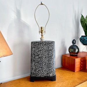 Puede incluir: Lámpara de mesa rectangular con un diseño de estampado de cebra en blanco y negro. La lámpara tiene un cuello color latón y una base blanca. La lámpara está sobre una superficie de madera.