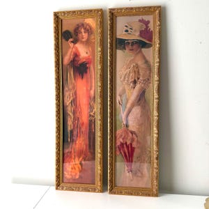 May include: Two vertical rectangular wall art prints in ornate gold frames. The left print depicts a person in a flowing orange gown holding a black fan. The right print shows a person in a light-colored dress and hat holding a pink parasol.