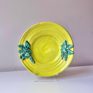 Puede incluir: Un plato de cerámica amarillo brillante con un borde acanalado y dos grupos de hojas verdes. El plato se exhibe sobre un soporte transparente sobre un fondo neutro. El diseño del plato es simple pero elegante.