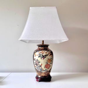 Puede incluir: Lámpara de mesa decorativa de cerámica con pantalla blanca. La base de la lámpara presenta un diseño floral y de pájaros detallado en tonos crema, marrón y dorado. La base se asienta sobre un soporte de madera oscura, creando un elegante acento de decoración del hogar.