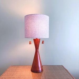 May include: A table lamp with a cylindrical, textured light gray fabric shade and a smooth, hourglass-shaped wooden base. The lamp features two pull chains with small wooden pendants hanging from beneath the shade to control the light.
