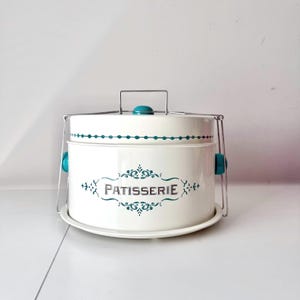 May include: A white metal cake carrier with a lid and a handle. The carrier has teal accents and the word "PATISSERIE" in teal script. The carrier sits on a white plate and has a metal handle.