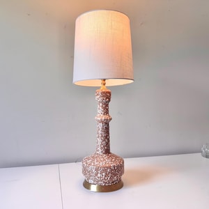 Puede incluir: Lámpara de mesa de estilo vintage con base de cerámica marrón moteada y pantalla de lino crema. La base tiene un borde de metal dorado. La lámpara está encendida, emitiendo un brillo cálido.