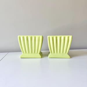 Puede incluir: Dos macetas de cerámica verde claro a juego. Cada maceta tiene una base rectangular y un cuerpo acampanado y acanalado. Las macetas son de un color verde pastel suave y están sobre un fondo blanco.