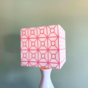 Könnte beinhalten: Ein quadratischer Lampenschirm mit weißem Hintergrund und einem sich wiederholenden geometrischen Muster aus leuchtend rosa Linien und Kreisen. Der Schirm ist auf einem glatten, weißen, konischen Lampenfuß montiert, der nach unten verläuft.