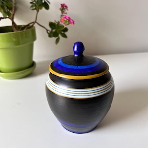 May include: A black ceramic jar with a lid, decorated with blue and gold stripes. The lid has a blue top.
