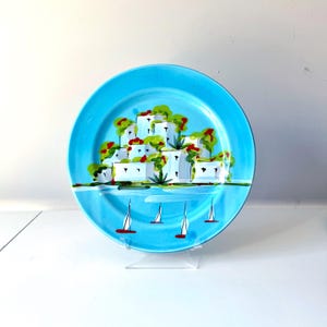 May include: A round, turquoise ceramic plate with a hand-painted coastal scene. The design features white buildings with green trees and red accents, along with white sailboats on the water. The plate is displayed on a clear stand.
