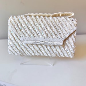 Puede incluir: Un bolso de mano blanco con cuentas, con solapa y asa pequeña. El bolso rectangular está cubierto de pequeñas lentejuelas y cuentas iridiscentes, creando un efecto texturizado y brillante. El diseño es clásico y elegante.