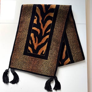 May include: A rectangular table runner featuring a central panel with a black and gold tiger stripe pattern, bordered by a leopard print section in shades of brown and black. The runner is finished with black fabric trim and four black tassels.