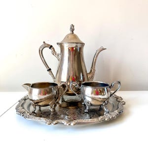 Puede incluir: Un juego de té plateado sobre una bandeja decorativa. El juego incluye una tetera, una crema y un azucarero. La tetera tiene un asa curva y un pico. La bandeja tiene un diseño floral alrededor de los bordes.