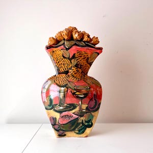 Puede incluir: Un jarrón de cerámica pintado a mano con un diseño único. El jarrón presenta una combinación de colores rojo y amarillo con ilustraciones de frutas, copas de vino y un arreglo floral en la parte superior. El jarrón mide unos 30 cm de alto.