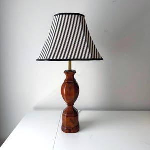 Puede incluir: Una lámpara de mesa de madera con una pantalla a rayas. La base es de color marrón oscuro con un diseño tallado. La pantalla es blanca con rayas verticales negras y un ribete negro.