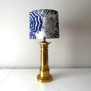 Puede incluir: Una lámpara de mesa dorada con base cilíndrica y pantalla. La pantalla presenta un diseño de estampado de cebra azul y blanco. La lámpara está sobre una superficie blanca.