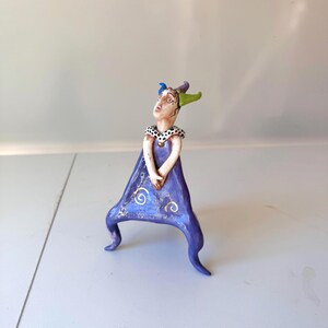 May include: A whimsical ceramic figurine of a person in a purple dress with gold accents. The figure has a black and white polka-dot collar and a hat with blue and green horns. The figurine is standing on two curved legs.