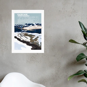 May include: A white and gray airplane flying over a snowy mountain range. The airplane has the text "KASE/ASE PITKIN CO./SARDY" on the side.