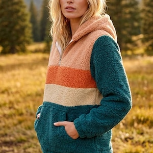 Puede incluir: Una sudadera con capucha de forro polar sherpa de bloques de color con cierre de media cremallera y bolsillo canguro frontal. La prenda presenta rayas horizontales en tonos melocotón, naranja y crema, con mangas y cuerpo inferior en azul cerceta.