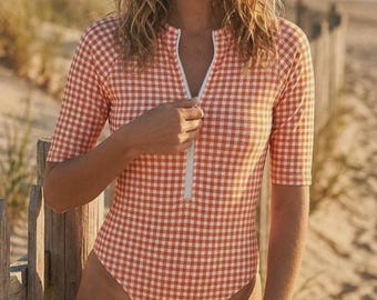 May include: A one-piece swimsuit with a red and white gingham pattern. The swimsuit has short sleeves and a white zipper down the front. The model is standing on a beach near a wooden fence.