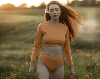 May include: An orange long-sleeved crop top and high-waisted bottoms set. The model is in a field of green grass and red flowers, with a flower in their hair. The background is a soft focus of a sunset.